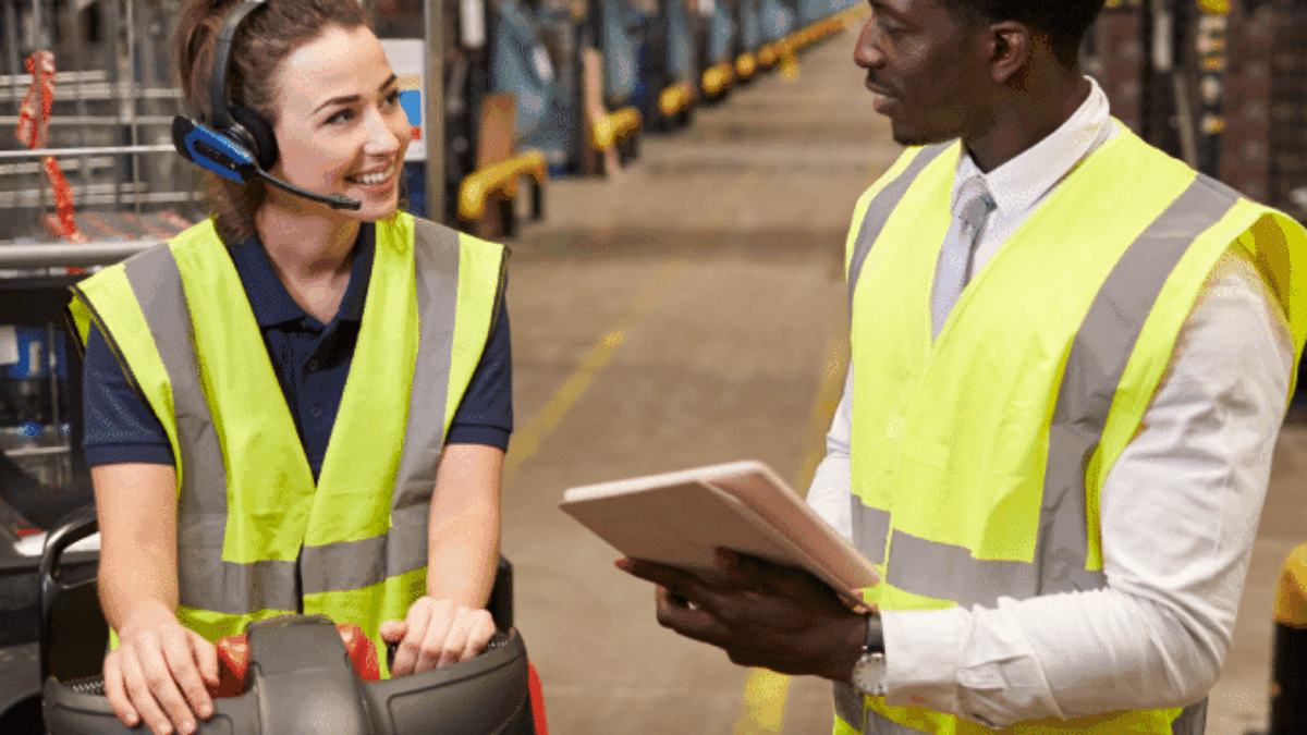 A woman wearing a headset and high-visibility vest sits on a forklift, smiling and talking to a man in a safety vest holding a tablet in a large warehouse.