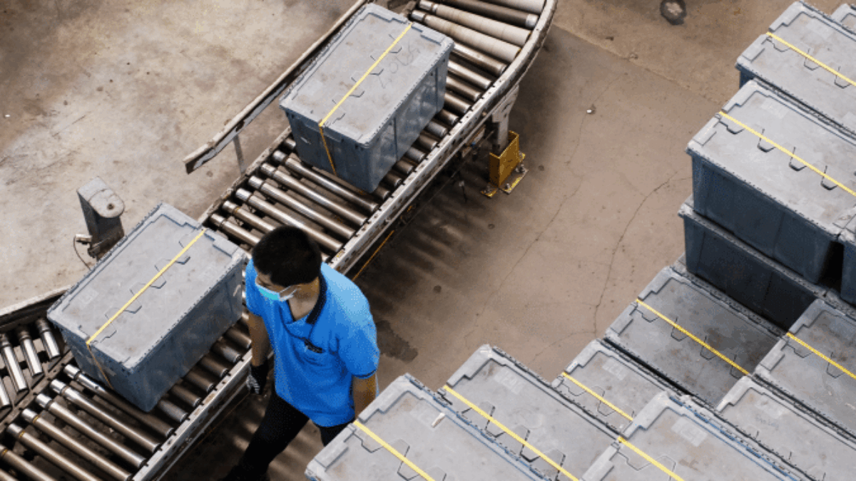 A worker in a blue shirt and mask walks next to a conveyor belt carrying gray storage bins inside an industrial warehouse. More gray bins are stacked nearby.