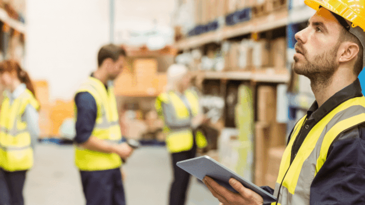 A warehouse worker in a yellow hard hat and safety vest holds a tablet and looks upward, with shelves of boxes in the background and other workers inspecting inventory.