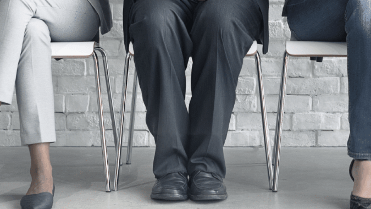 Three people in business attire sit on chairs against a white brick wall, each holding a smartphone or tablet, with only their lower bodies visible.