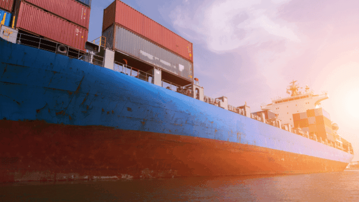 A large blue cargo ship loaded with stacked shipping containers sails on calm water under a clear sky, with the sun setting in the background.