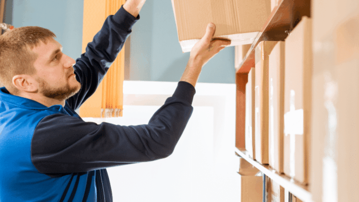 A man in a blue vest is reaching up to place or retrieve a cardboard box from a high shelf in a storage area or warehouse with several other boxes on shelves.