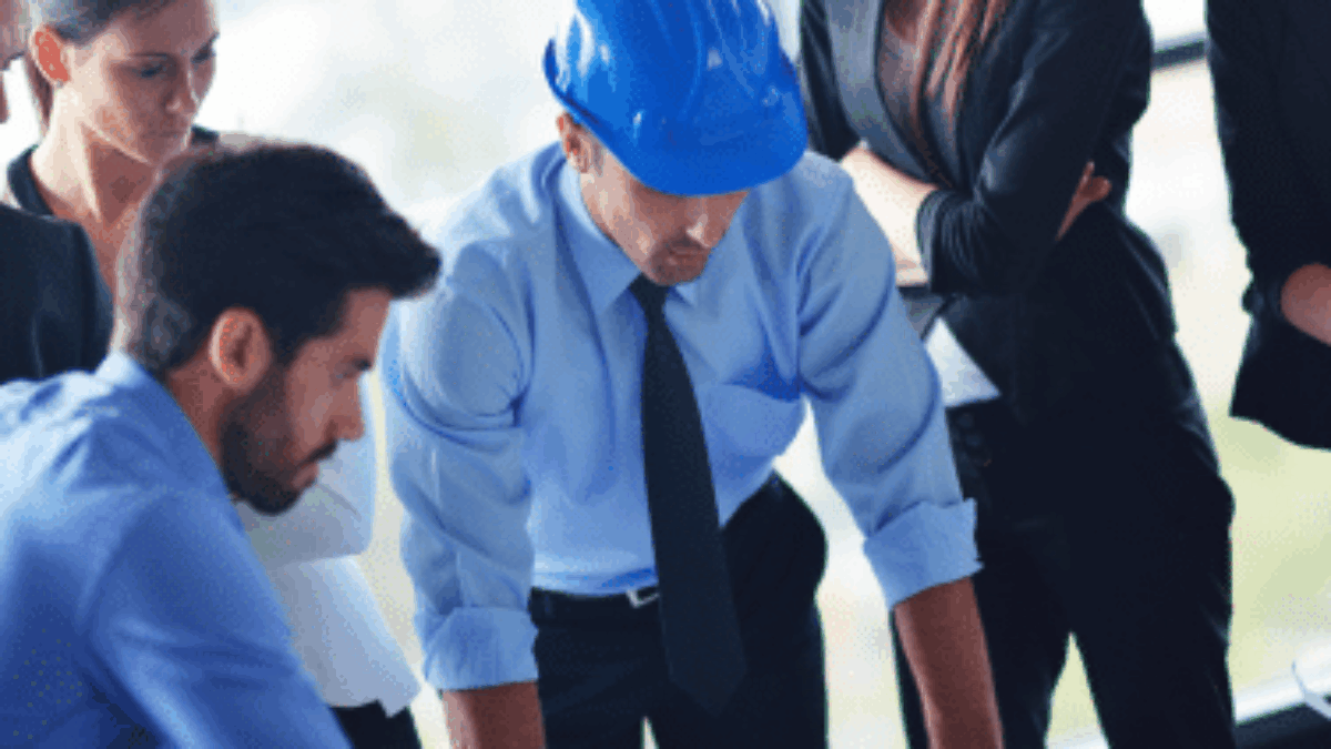A group of professionals, including a person wearing a blue hard hat, closely examining blueprints on a table in a modern office setting.