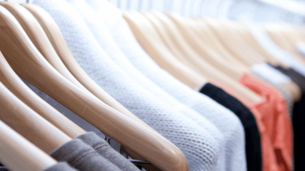 A row of wooden hangers holding various sweaters and shirts in neutral and warm colors on a clothing rack in a store or closet.
