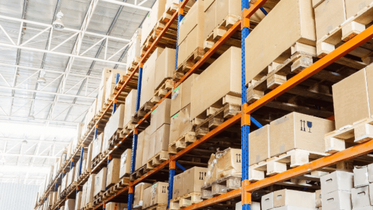 Large warehouse interior with tall blue and orange metal shelves stacked with brown cardboard boxes and wooden pallets, extending into the distance under a high ceiling with exposed beams.