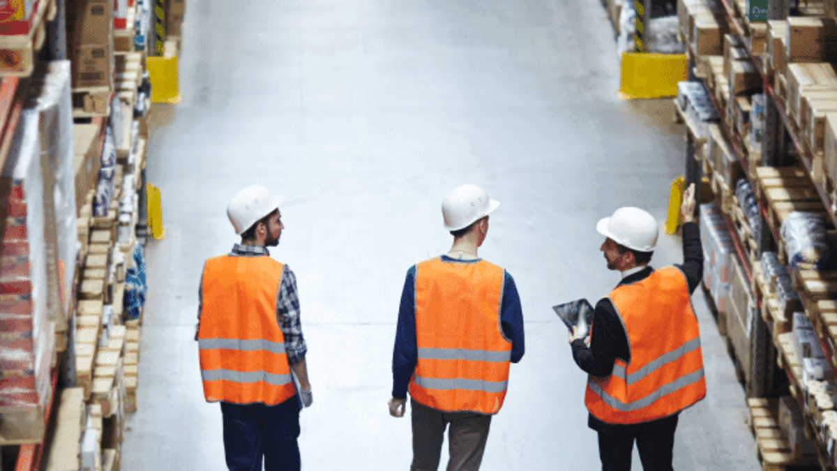 Three workers wearing orange safety vests and white hard hats walk down an aisle in a warehouse lined with shelves full of boxes and products. One worker gestures towards the shelves while another holds a clipboard.