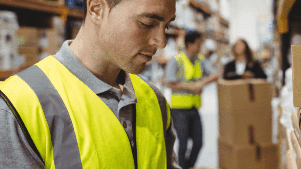 A warehouse worker in a yellow high-visibility vest scans a box with a handheld device, with shelves of packages and other workers visible in the background.