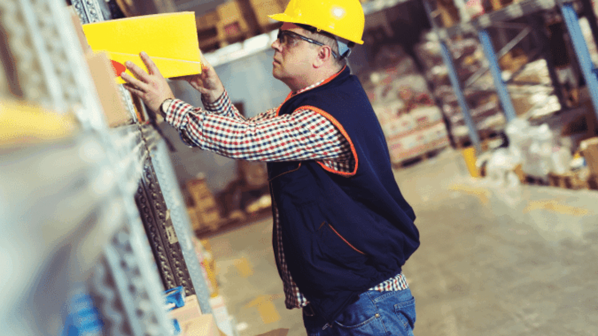 A warehouse worker in a yellow hard hat and safety vest stands by metal shelves, reaching to organize or retrieve a yellow box. The warehouse is filled with boxes and goods on racks.