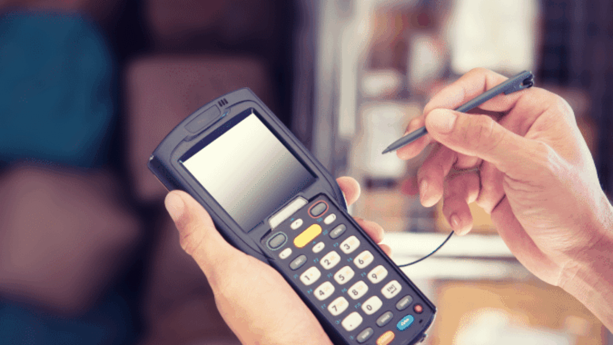 A person uses a stylus to operate a handheld electronic device with a keypad and screen, likely for inventory or data entry, in a warehouse or storage setting.
