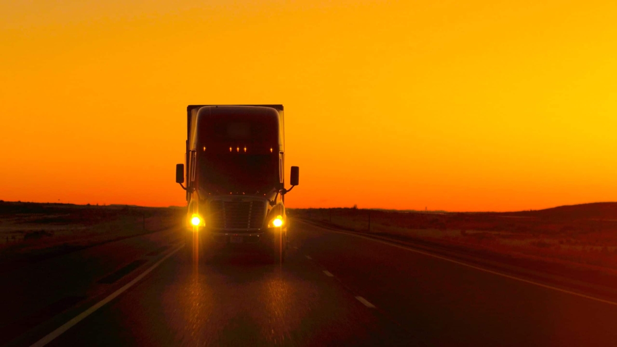 A large semi-truck with headlights on drives down an empty highway at sunset, silhouetted against a vibrant orange sky.