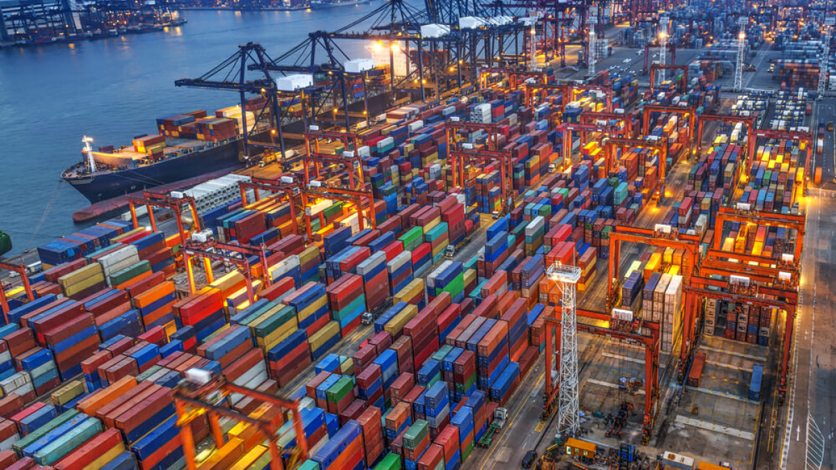 An aerial view of a busy shipping port at dusk, filled with colorful stacked shipping containers, large cranes, and cargo ships docked by the water, with city buildings visible in the background.
