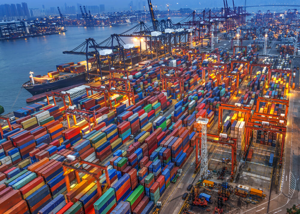An aerial view of a busy shipping port at dusk, filled with colorful stacked shipping containers, large cranes, and cargo ships docked by the water, with city buildings visible in the background.