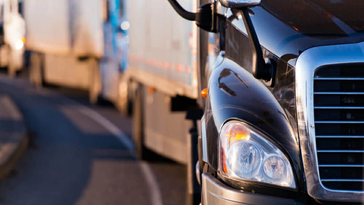 Close-up of the front of a black semi-truck driving on a road, with several other trucks visible in a line behind it, curving along the road.