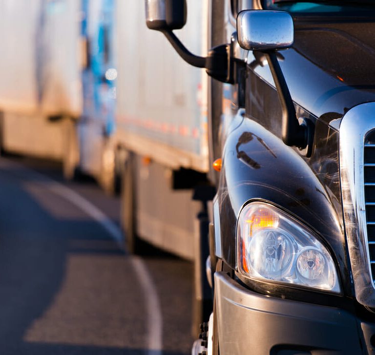 Close-up of the front of a black semi-truck driving on a road, with several other trucks visible in a line behind it, curving along the road.