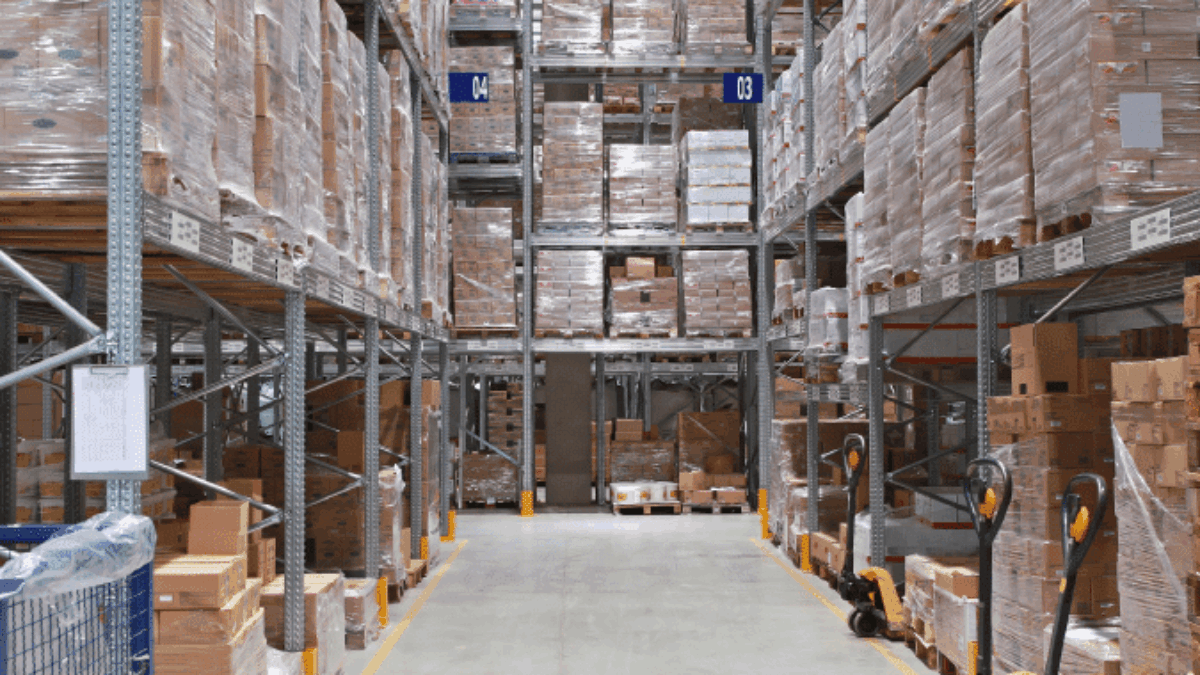A warehouse aisle with tall shelves stacked with pallets of boxed goods. Pallet jacks are visible on the right, and the floor is clear, allowing access for transport and movement.