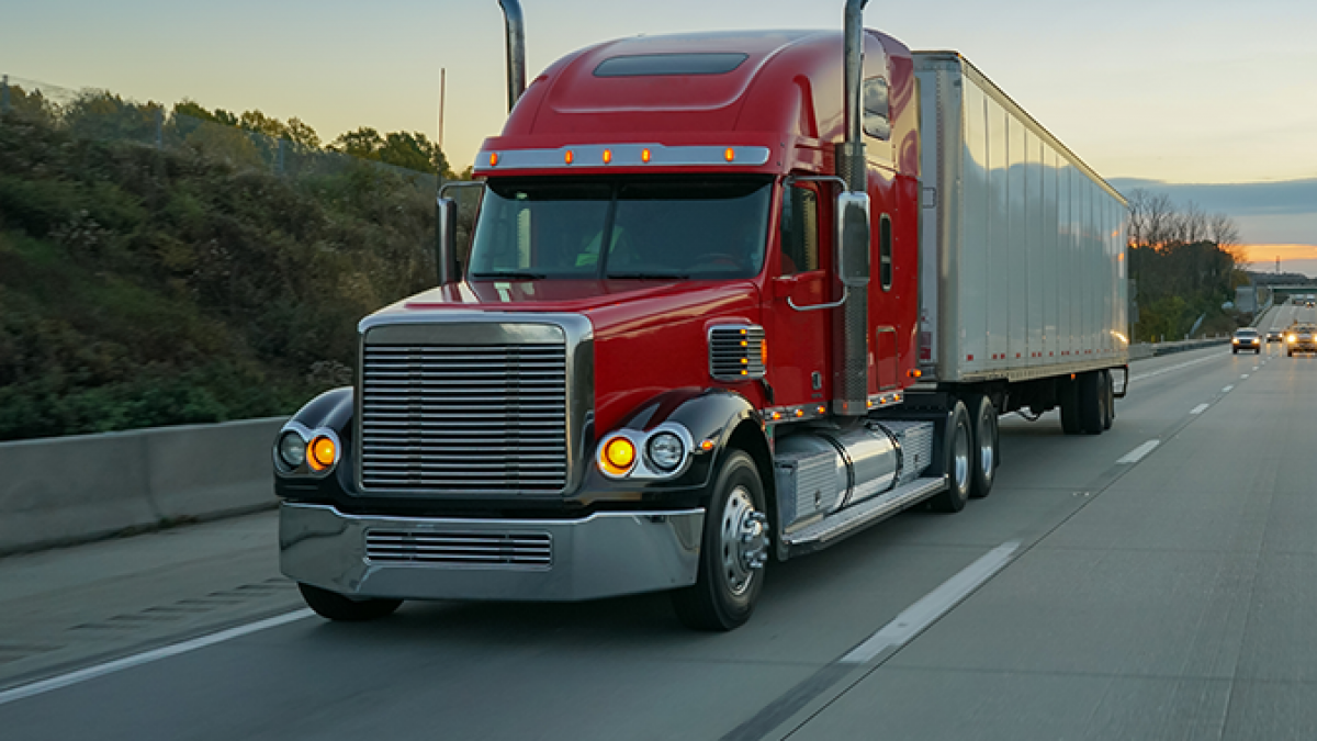 A red semi-truck with a large silver trailer drives on a highway at dusk, passing grassy roadside and trees under a clear blue sky.