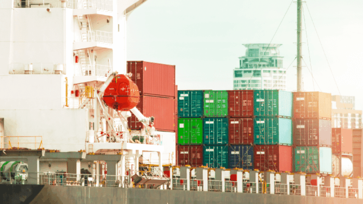 A large cargo ship loaded with colorful shipping containers docked at a port, with modern buildings visible in the background under a bright sky.