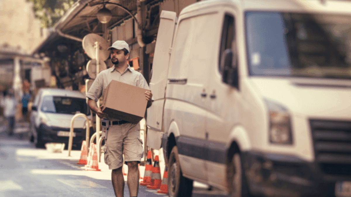 A delivery worker in a uniform and cap carries a cardboard box on a city street, walking near a white van parked at the curb with traffic cones nearby. Trees and other vehicles are in the background.