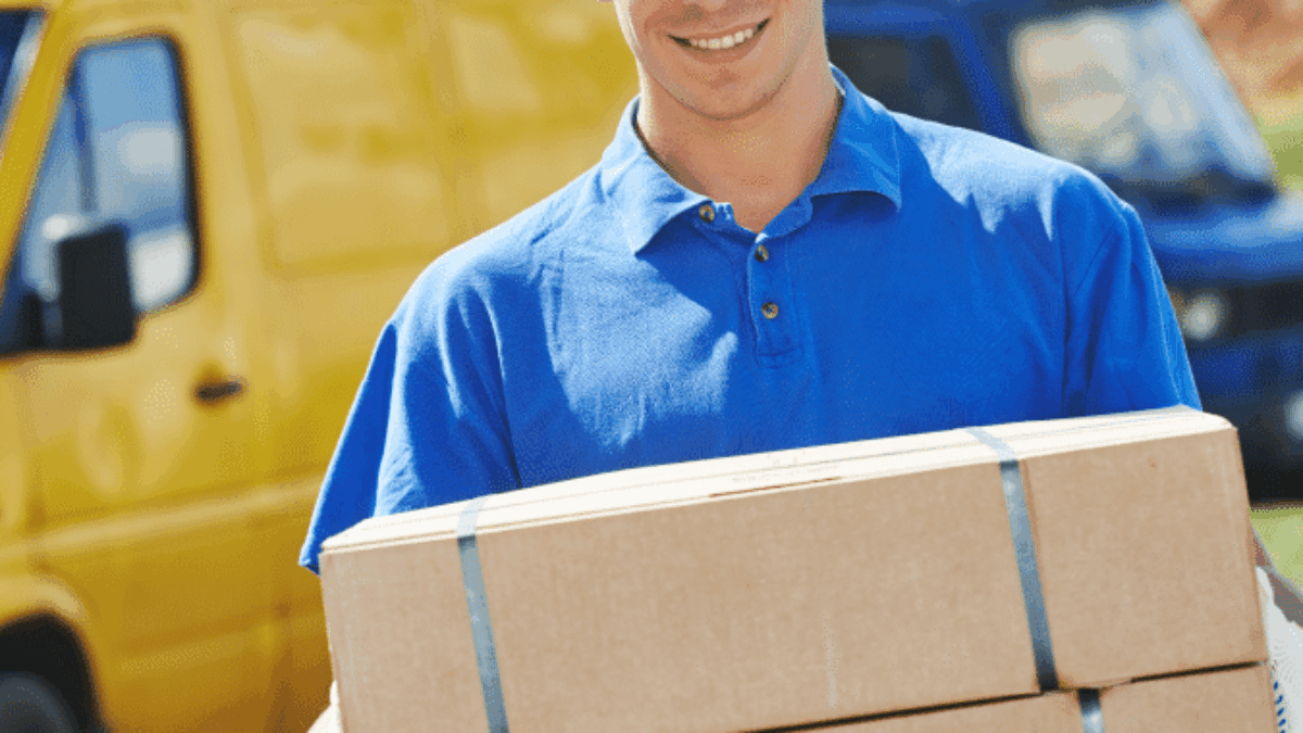 A smiling delivery person in a blue uniform and cap holds two cardboard boxes. A yellow van and a blue van are parked in the background.
