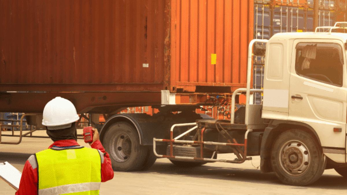 A worker in a reflective vest and hard hat inspects a large shipping container on a truck at a busy shipping yard with stacked containers in the background.