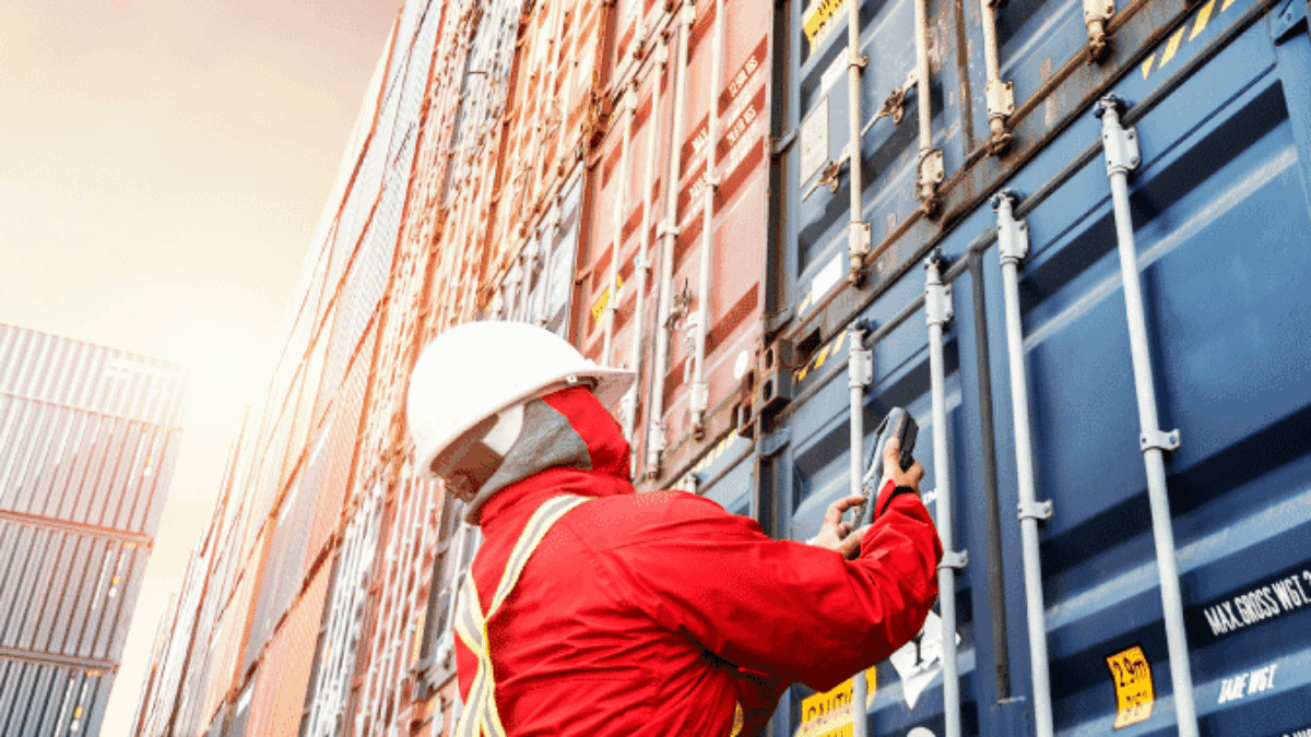 A worker in a red uniform and white hard hat inspects or secures stacked shipping containers outdoors, with sunlight shining in the background.