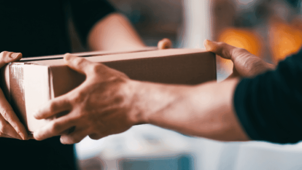 Two people exchanging a cardboard box indoors, with one person handing it over to the other; their faces are not visible.