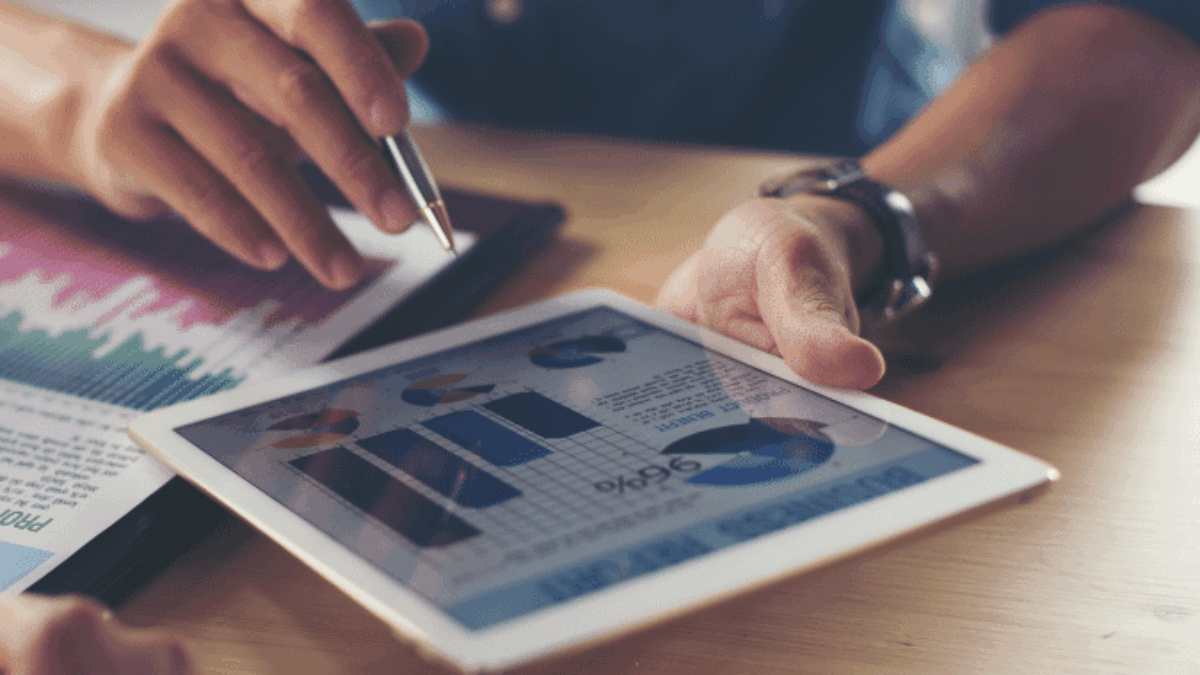 Two people discuss financial data at a wooden table. One holds a pen and points to a tablet displaying charts and graphs, while the other reviews printed documents with similar information.