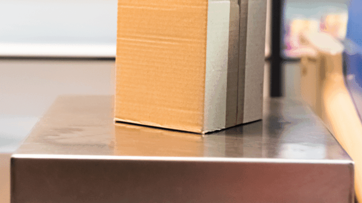 A small cardboard box sits on a metal platform or scale, possibly in a shipping or warehouse setting, with blurred shelves in the background.