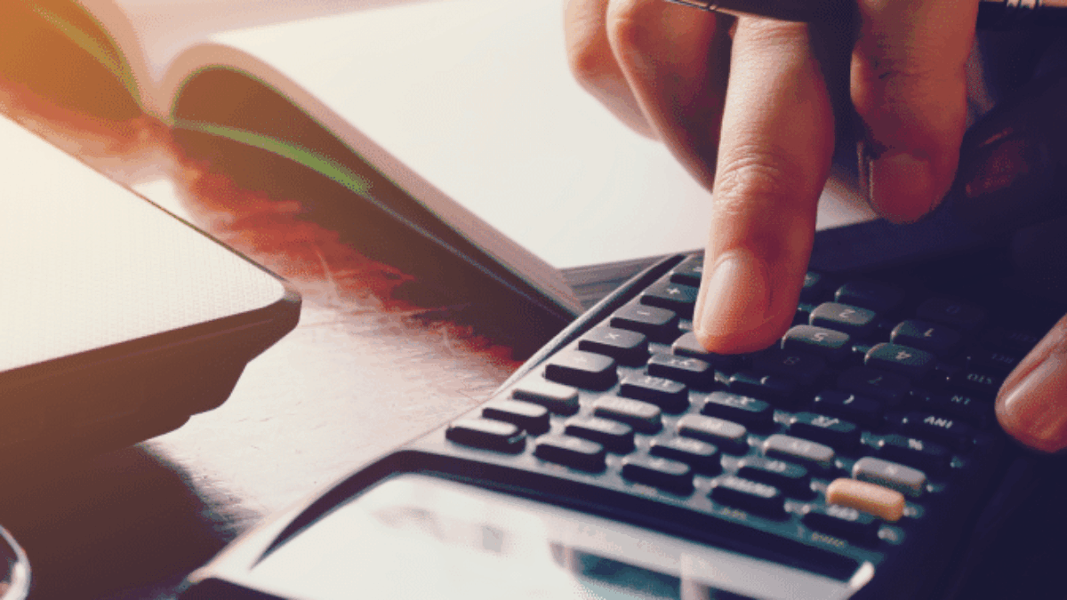 A person uses a calculator with one hand while holding a pen in the other, sitting at a desk with an open notebook, a laptop, and orange sticky notes nearby. Sunlight shines across the workspace.