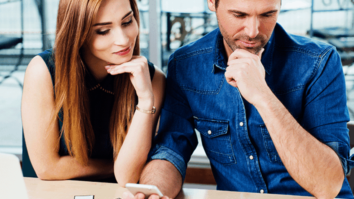 A man and woman sit at a desk, thoughtfully looking at a smartphone and colorful cards arranged on the table. A laptop and pens are nearby, suggesting a collaborative planning or brainstorming session.