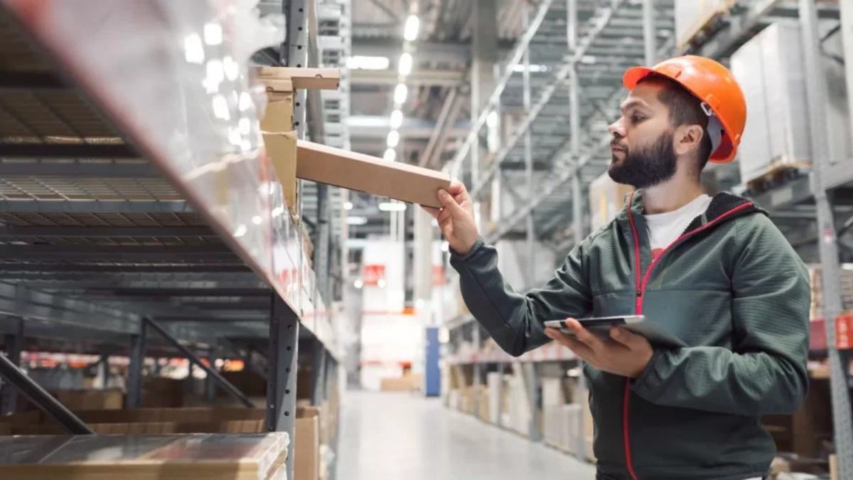A man wearing an orange hard hat and a green jacket stands in a warehouse, holding a tablet and placing a box on a shelf. The warehouse has tall shelves stocked with various items.