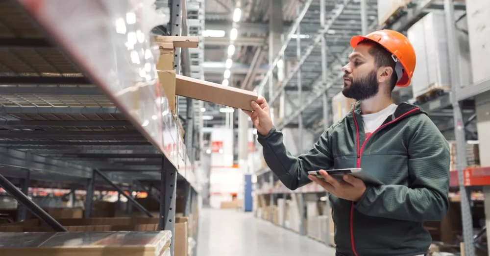 A man wearing an orange hard hat and a green jacket stands in a warehouse, holding a tablet and placing a box on a shelf. The warehouse has tall shelves stocked with various items.