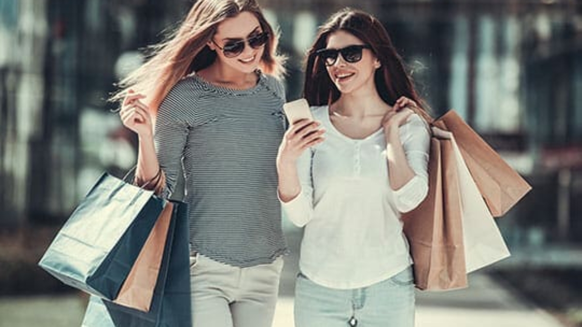 Two young women wearing sunglasses and casual clothes walk outdoors, smiling and holding shopping bags. One is looking at a smartphone while the other glances at the screen.