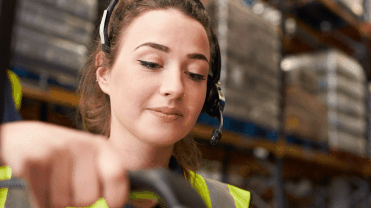 A woman wearing a headset and high-visibility vest scans a package with a handheld barcode scanner in a warehouse filled with shelves and boxes.