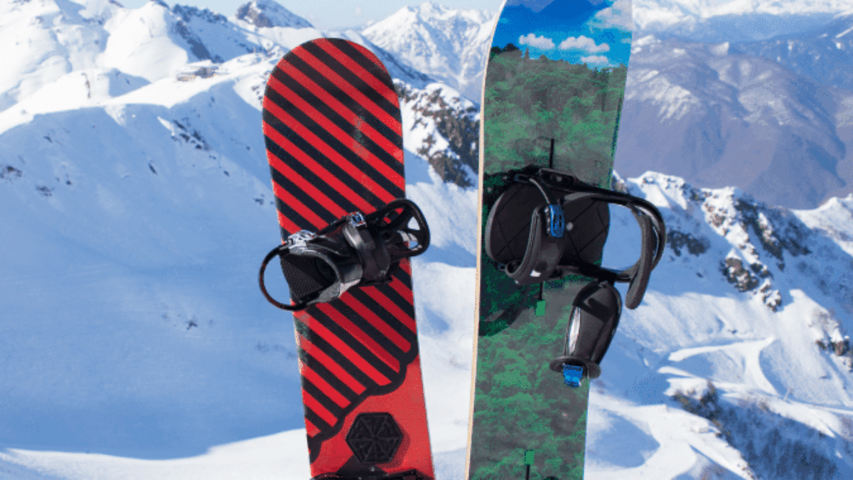Two snowboards, one red with black stripes and one with a green and blue nature design, standing upright in the snow on a mountain with snowy peaks in the background under a clear blue sky.