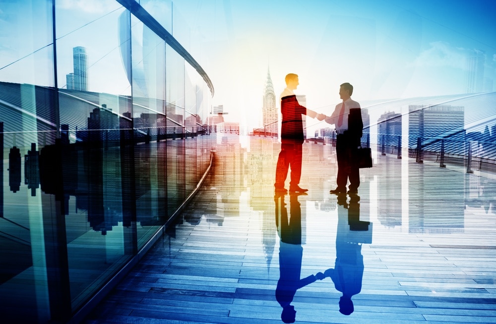 Two businesspeople shake hands on a modern glass walkway, with a city skyline and tall buildings visible in the background, and their reflections on the shiny floor.