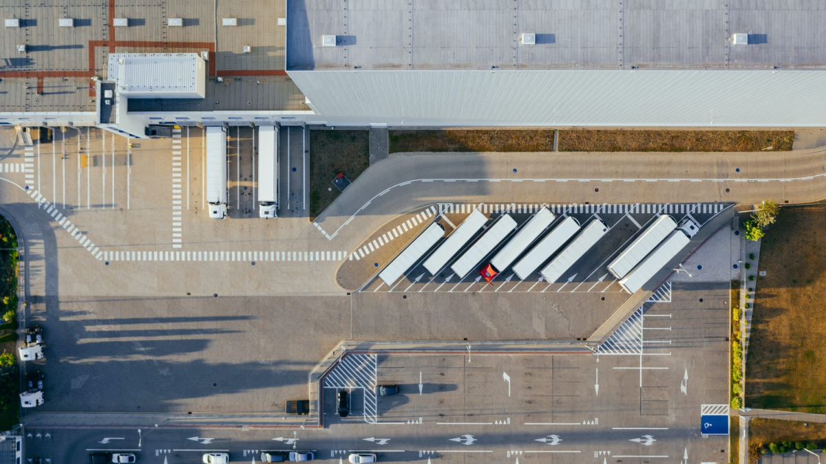 Aerial view of a warehouse with several trailers parked in loading bays, a few trucks at the docks, and a parking lot with cars in the foreground. The area is organized with marked lines and roads.