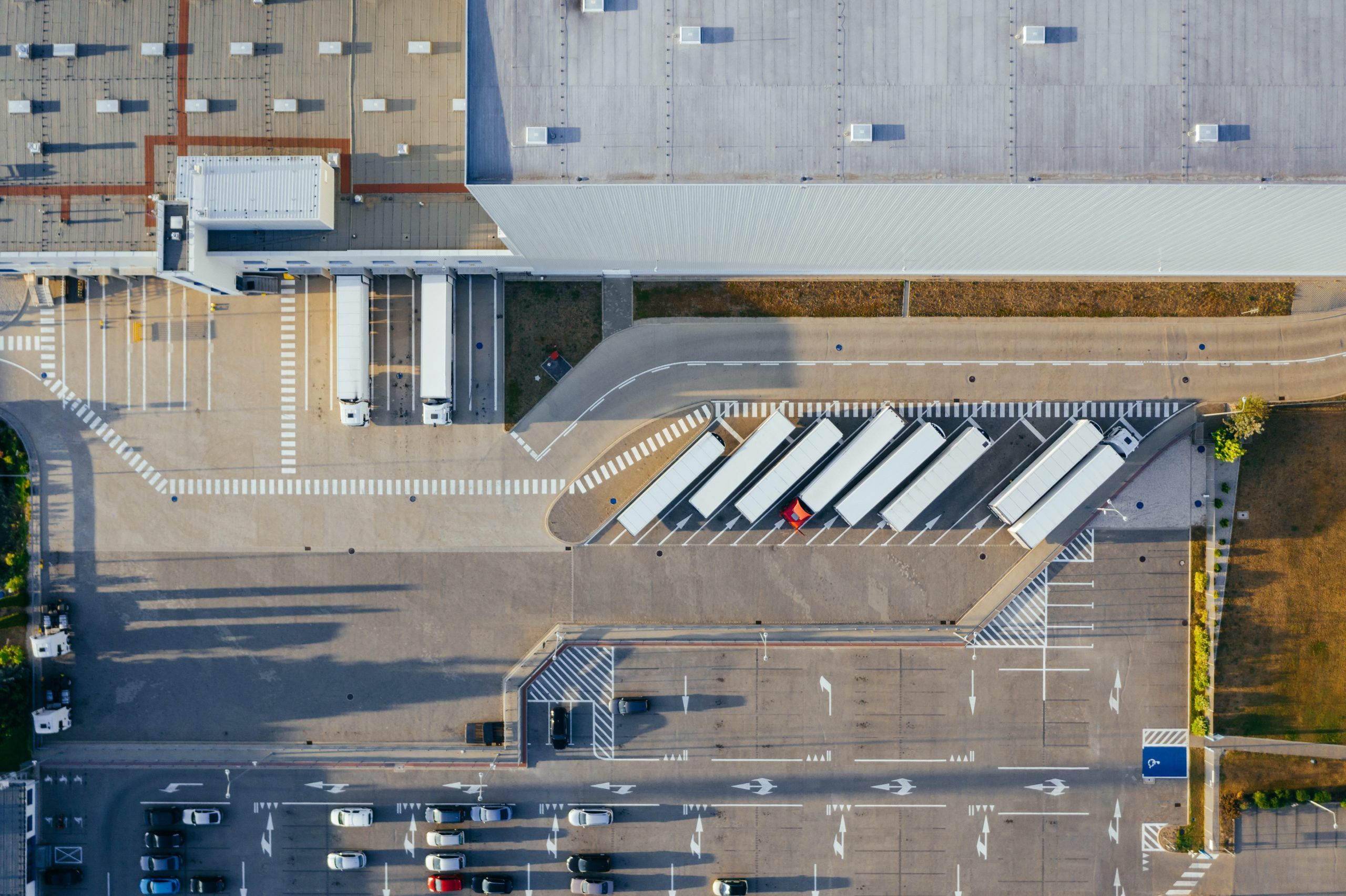 Aerial view of a warehouse with several trailers parked in loading bays, a few trucks at the docks, and a parking lot with cars in the foreground. The area is organized with marked lines and roads.