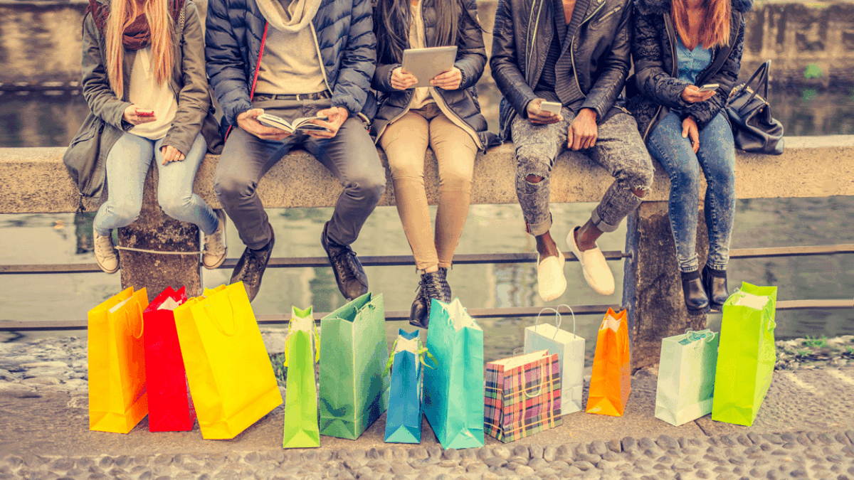 Five people sit on a stone bench outdoors, each with shopping bags of various colors and patterns placed in front of them. They are dressed in casual winter clothes and appear to be using their phones or reading.