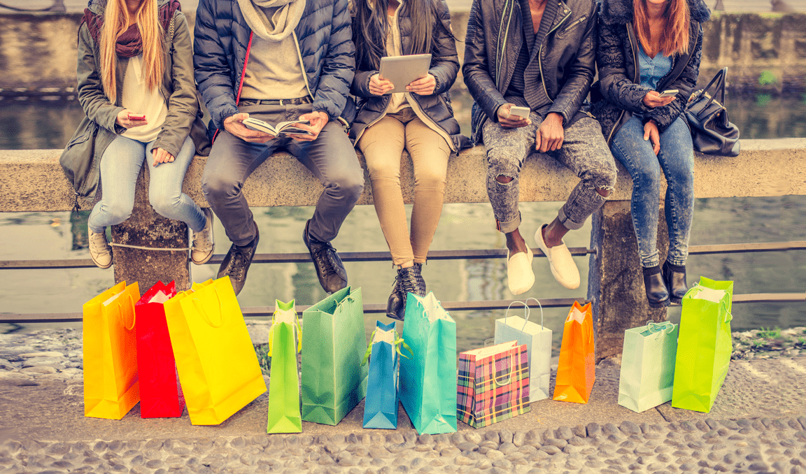 Five people sit on a stone bench outdoors, each with shopping bags of various colors and patterns placed in front of them. They are dressed in casual winter clothes and appear to be using their phones or reading.
