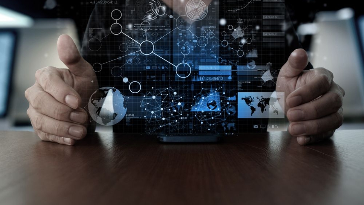 A person sits at a table with their hands around a digital interface displaying futuristic graphics, charts, world maps, and data visualizations, suggesting advanced technology and data analysis.