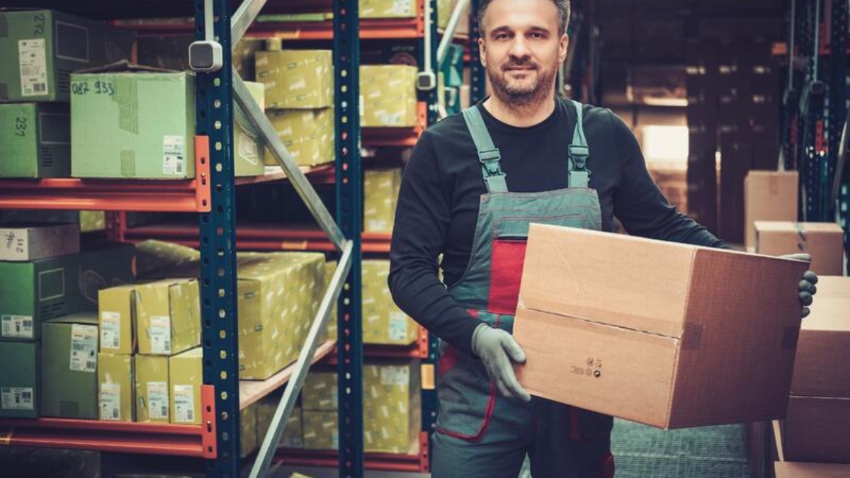 A warehouse worker in overalls and gloves holds a cardboard box, standing among shelves filled with packages and parcels. The storage area is organized and well-lit.