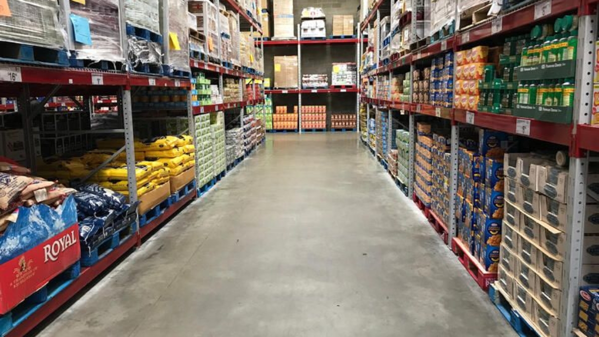A wide aisle in a warehouse-style grocery store with shelves stacked high with bulk food items and large boxes on both sides. The concrete floor is clean and there are no shoppers visible.