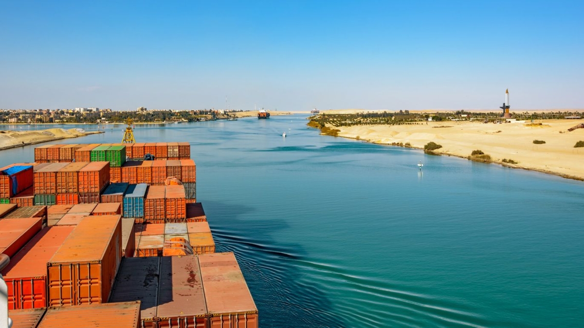 A cargo ship loaded with containers sails through the Suez Canal, bordered by sandy desert on one side and a distant cityscape on the other, under a clear blue sky.