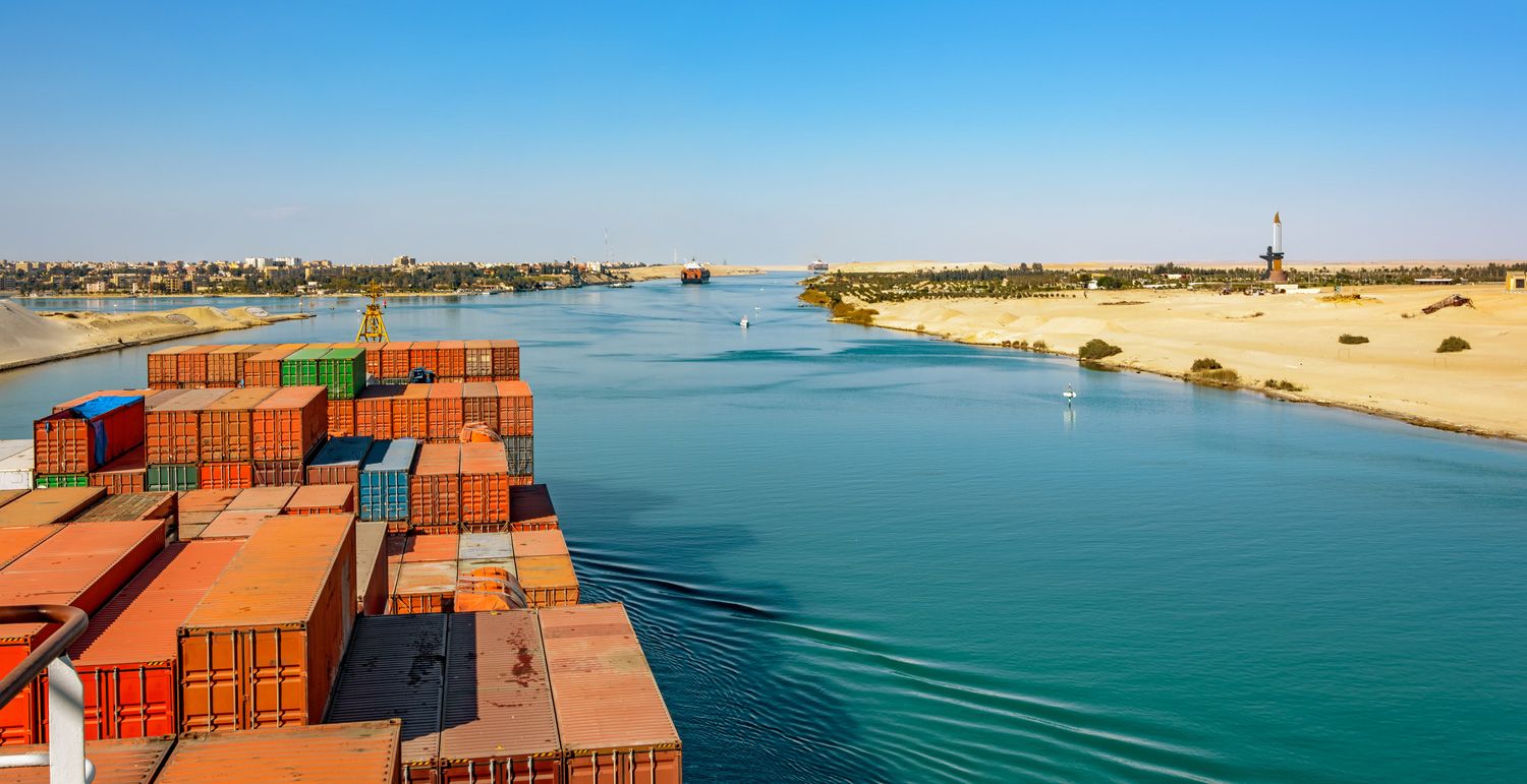 A cargo ship loaded with containers sails through the Suez Canal, bordered by sandy desert on one side and a distant cityscape on the other, under a clear blue sky.