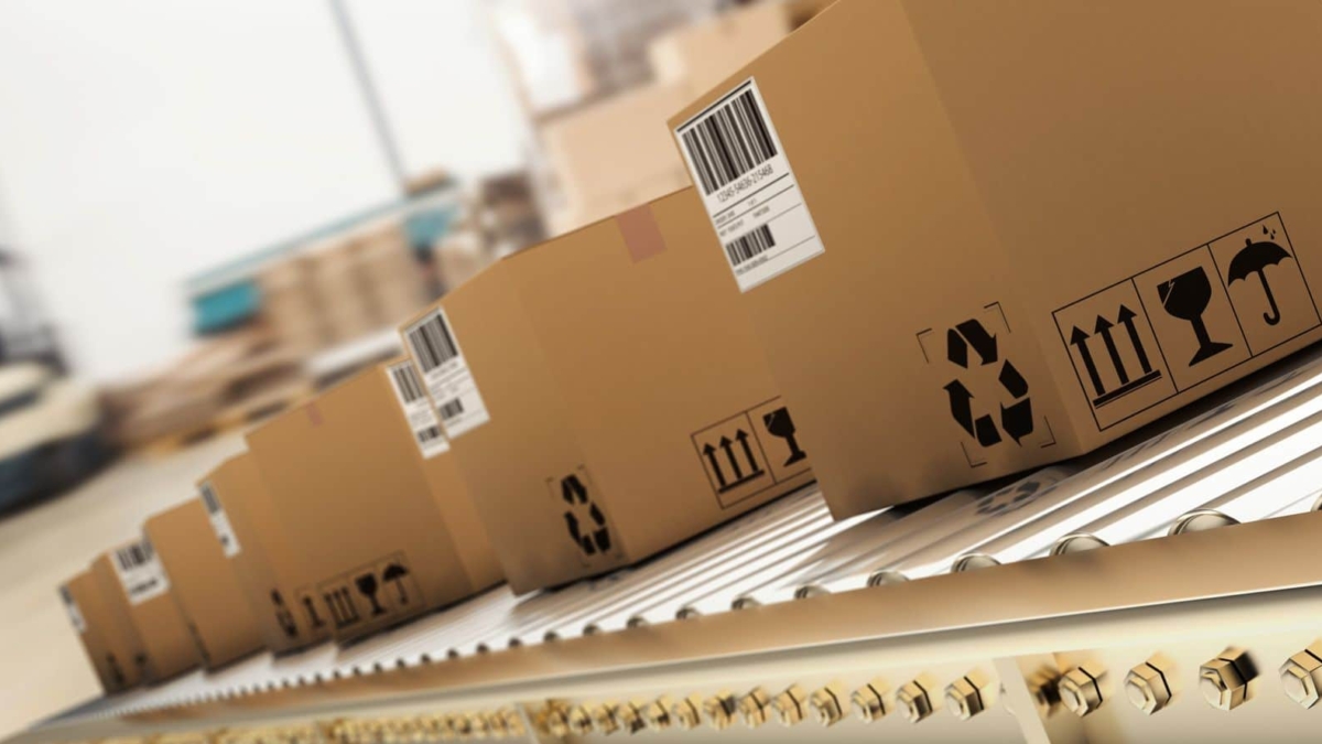 Several cardboard boxes with barcodes and shipping symbols move along a conveyor belt in a warehouse or distribution center, suggesting a shipping or logistics process.