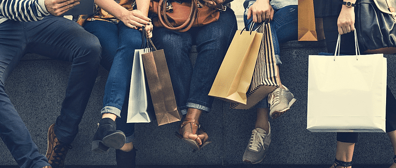 Picture1 copy Four people sitting side by side outdoors, holding multiple shopping bags. Only their legs, hands, and bags are visible, suggesting they have been shopping together.