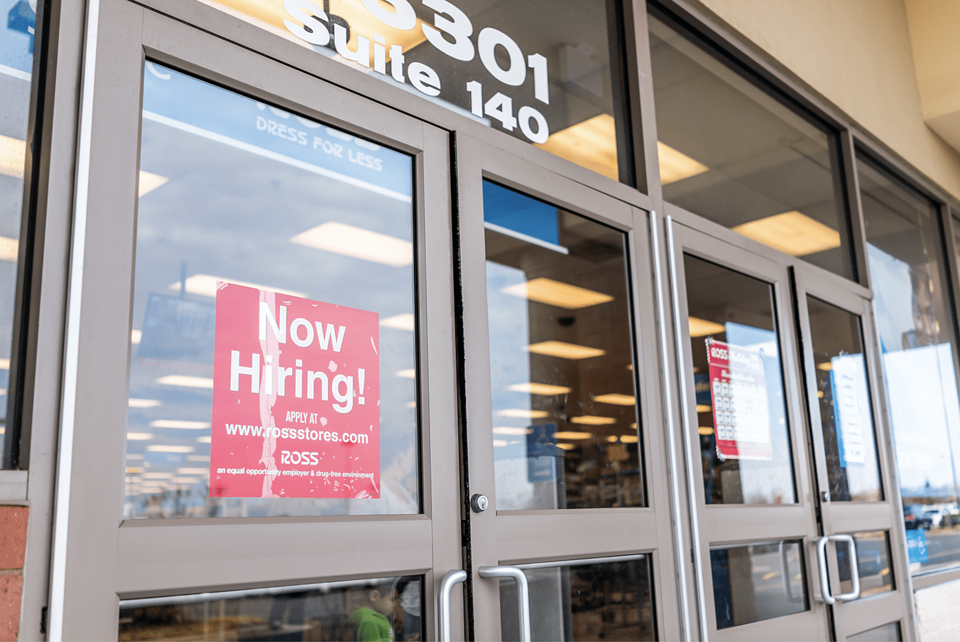 A glass storefront door displays a red “Now Hiring!” sign with application details for Ross. The store’s interior is visible through the windows, showing fluorescent lights and merchandise.