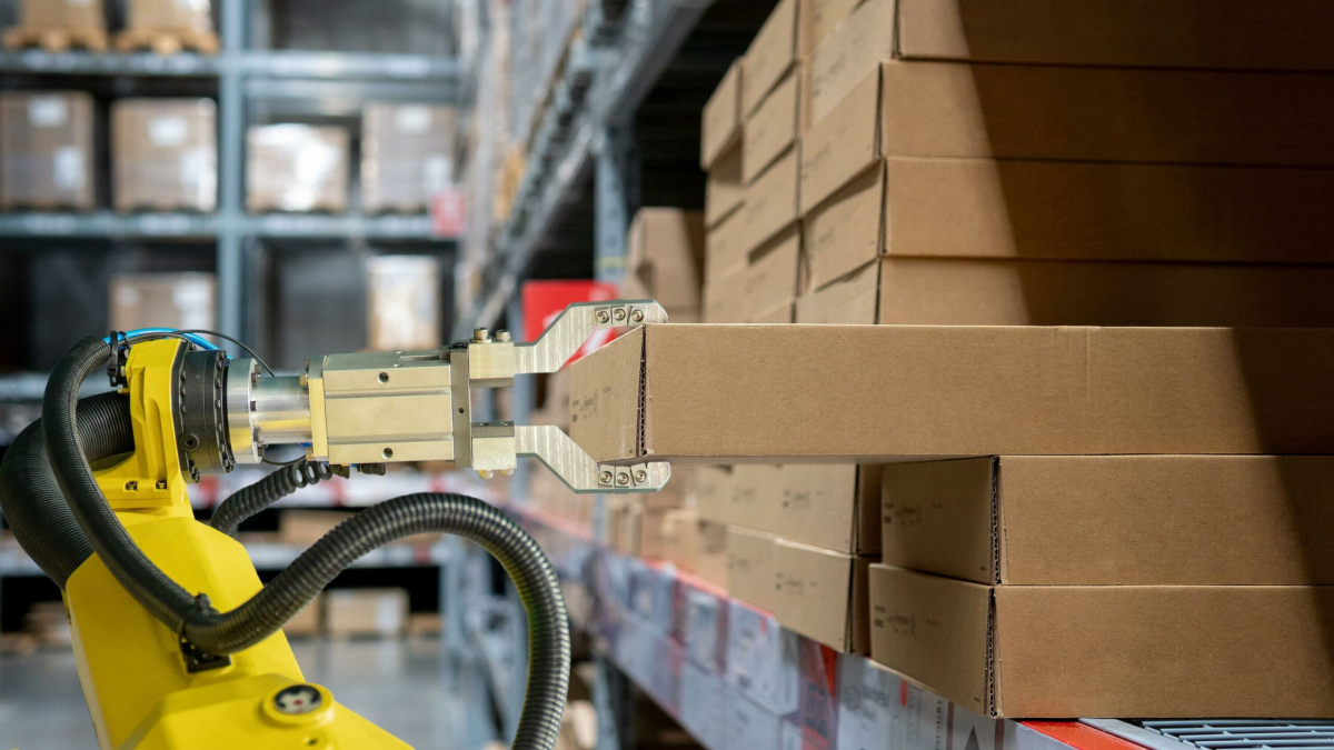 A robotic arm with a metal gripper is picking up a cardboard box from a shelf in a warehouse filled with stacked brown boxes.