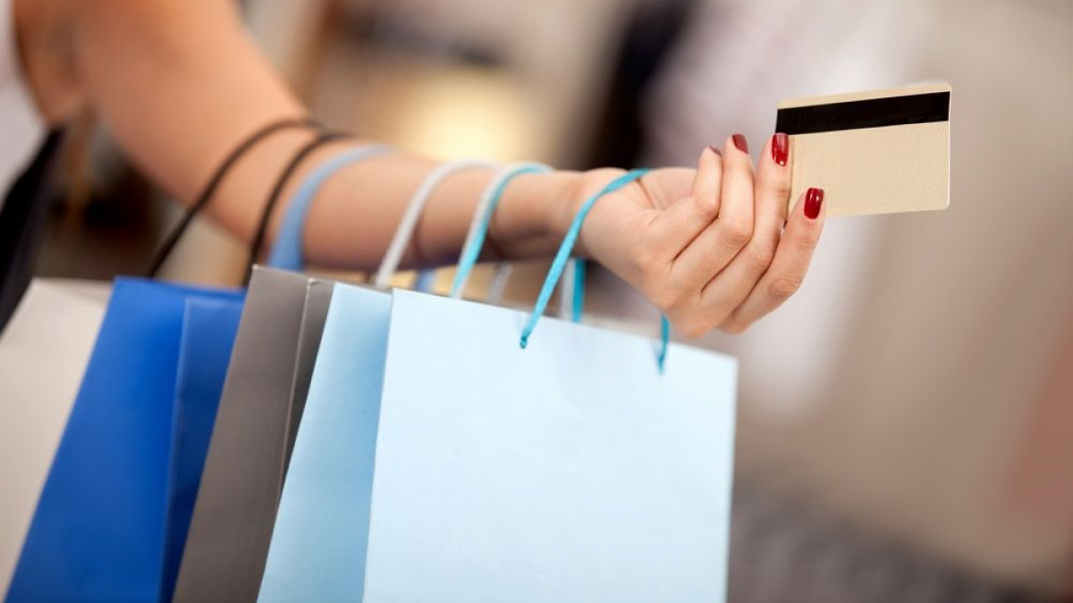 A person with red-painted nails holds several shopping bags and a credit card, suggesting a shopping activity. The background is blurred, keeping the focus on the hand, bags, and card.